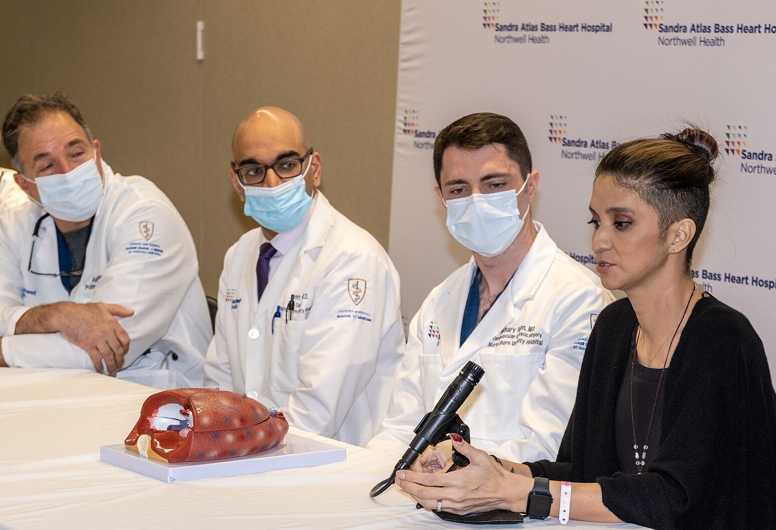 A woman wearing a black shirt speaks into a microphone while four doctors wearing white lab coats and face masks look on.
